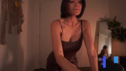 Media: Video of a slim, light-skinned woman with short black hair in a tight, dark brown leather dress, standing in a dimly lit, cozy room with a large mirror and wall art.