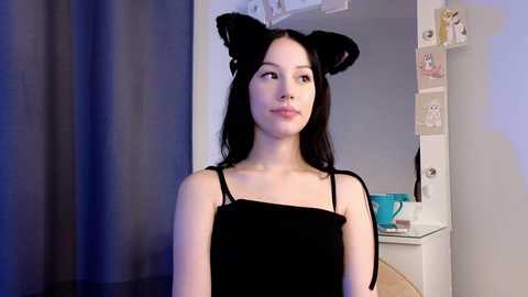 Media: Video of a young East Asian woman with long black hair, wearing black cat ears and a black velvet dress, standing indoors with gray curtains and white shelves in the background.