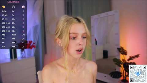 Media: Video of a nude, blonde, fair-skinned woman with a soft, natural face, in a dimly lit, artistic studio setting with abstract artwork, potted plants, and a laptop displaying a video recording interface.