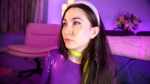 Media: Video of a young woman with long dark hair and fair skin, wearing a shiny purple top and yellow scarf, sitting in a modern living room with pink and purple lighting.