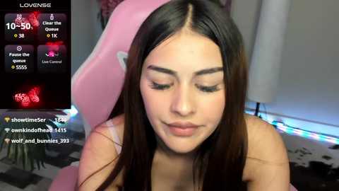 Media: A video of a young woman with long, straight brown hair, light skin, and closed eyes, seated on a pink gaming chair in a dimly lit room.