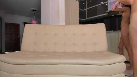 Media: Video of a beige tufted sofa in a modern, minimalist living room with wooden doors, a white column, and a person partially visible on the right.