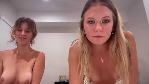 Media: Video of two topless women in a minimalist room: one with shoulder-length blonde hair, large breasts, and a cross necklace; the other with wavy brown hair, smaller breasts, and wearing earrings.