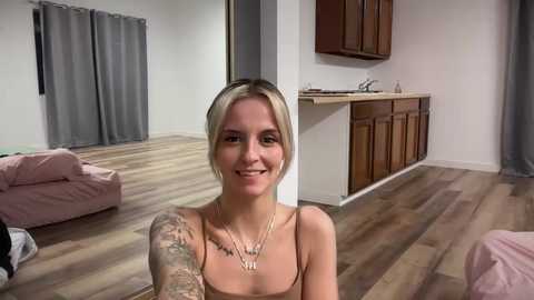 Media: Video of a blonde woman with a tattooed arm, smiling, sitting in a modern, light-colored living room with wooden floors and gray curtains.