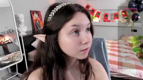 Media: Video of a young woman with fair skin and long brown hair, wearing a silver headband and elf ears, sitting on a bed with a striped blanket. The room has a white wall with a shelf displaying Halloween decorations.