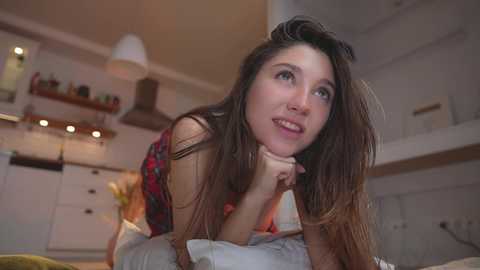 Media: Video of a young woman with long brown hair, wearing a red plaid top, leaning on a bed in a cozy, dimly-lit bedroom with white walls and wooden shelves.