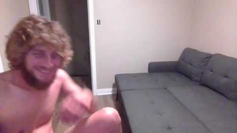 Media: Video of a nude man with curly hair, smiling, sitting on a grey sectional sofa in a beige-walled room.