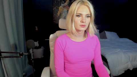 Media: Video of a blonde woman in a bright pink long-sleeved top, seated in a dimly lit room with a white chair, bed, and dark walls.