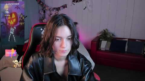 Media: Video of a young woman with shoulder-length dark hair, wearing a black leather jacket, sitting in a red gaming chair. Background features a Halloween-themed wall, decorations, and a couch.