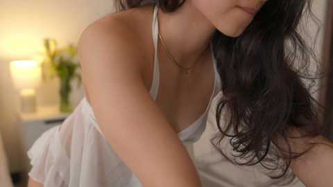 Media: Video of a woman with long dark hair in a white, sheer nightgown, leaning over a bed with a warm lamp in the background.