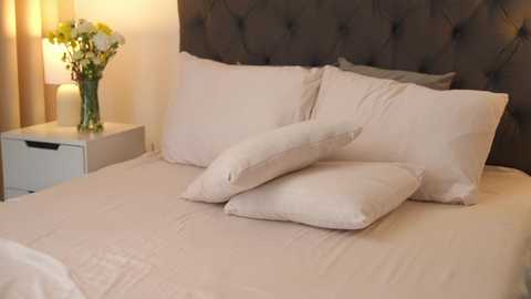 Media: Video of a tidy, beige-tiled bedroom with a tufted dark grey headboard, a cream bedspread, two white pillows, and a matching white nightstand with a white lamp and yellow flowers.