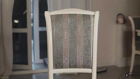 Media: Video of a white wooden chair with a glass panel back, positioned indoors near a large window with white curtains. Background features a minimalist room with a white wall and a small decorative item on a shelf.