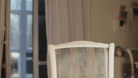 Media: Video of a vintage, floral-patterned upholstered chair with a white frame, set against a softly lit, cozy room with white curtains and a window, and decorative items on a side table.