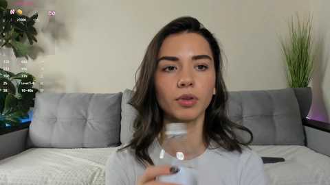 Media: Video of a young woman with long, dark hair, fair skin, and light makeup, wearing a white shirt, sitting on a gray couch. Background includes a potted plant, a green vase, and a TV screen showing a live feed.