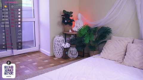 Media: Video of a serene, minimalist bedroom with white bedding, white drapes, and a wooden bedside table adorned with green plants and white ceramic vases.