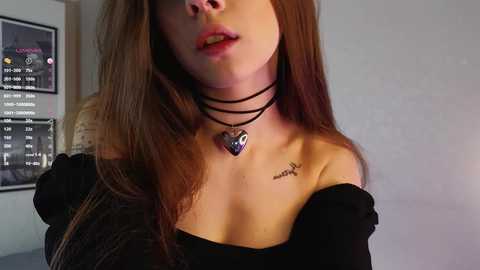 Media: Video of a young woman with long brown hair, wearing a black off-shoulder top, black choker necklace, and a heart pendant. She has a tattoo on her left shoulder. Background features a framed calendar and wall.