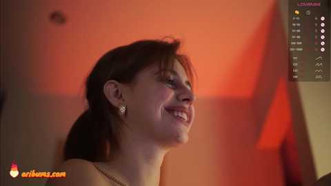 Media: Video of a smiling woman with shoulder-length brown hair, wearing a strapless top, set against an orange backdrop with a website watermark.