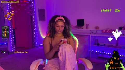 Media: Video of a young Black woman with curly hair, wearing a white headband, playing a video game in a purple-lit room with Halloween decorations and a Batman poster.