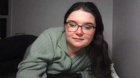 Media: Video of a fair-skinned woman with long black hair and glasses, wearing a green zip-up hoodie, leaning forward, smiling slightly. Background features a plain, light-colored wall and a black chair.