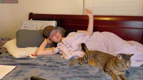 Media: A video of a young girl with light skin and short blonde hair, lying in bed with a gray cap, pink blanket, and a gray cat.