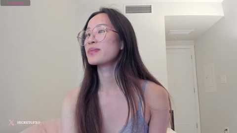 Media: Video of an Asian woman with long black hair, wearing glasses and a gray tank top, standing in a minimalist white room with doors and an air conditioner vent.