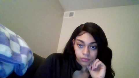 Media: A young woman with long, straight black hair and fair skin rests her chin on her hand, wearing a black top. She gazes pensively against a beige wall and a blue-and-white checkered pillow in the background.