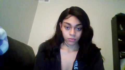 Media: Video of a young woman with long, straight black hair, wearing a black jacket, looking down, in a dimly lit room with a white wall and a black shelf in the background.