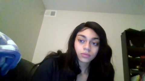 Media: A video of a young woman with long, straight black hair, fair skin, and dark eyes, wearing a black top, sitting in a dimly lit room with a white wall and a dark bookshelf in the background.