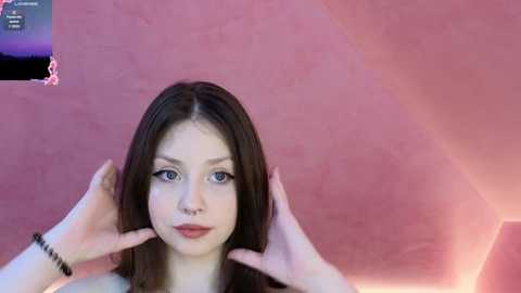 Media: A video of a young Caucasian woman with long brown hair, light skin, and blue eyes, wearing red lipstick, touching her ears. The background is a pink, textured wall with a lens flare.