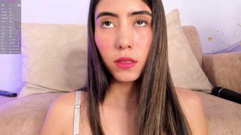 Media: Video of a young woman with long, straight brown hair, light skin, and pink lipstick, wearing a light-colored top, sitting on a beige couch in a softly lit room.