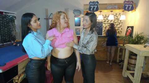 Media: Video of three women in a pool hall: one lifting her pink top, another in black pants, and the third in a striped shirt, smiling.