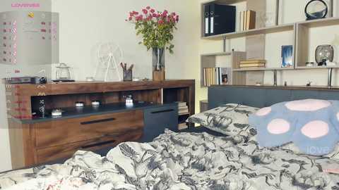 Media: Video of a modern bedroom with a wooden dresser, bookshelves, floral arrangement, polka-dot pillows, and a bed with a patterned duvet.