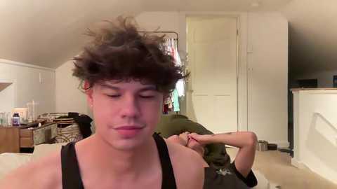 Media: Video of a young man with curly hair and a black tank top, smiling, sitting on a couch in a cluttered living room with a person in the background.