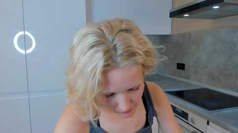 Media: Video of a blonde woman with wavy hair, wearing a gray tank top, focused on a sleek, modern kitchen with white cabinets, gray countertops, and a black stove.