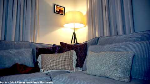 Media: Video of a cozy living room with a sectional sofa featuring various textured cushions in shades of gray and purple, illuminated by a warm-toned floor lamp. Soft, white curtains frame the space, adding to the serene ambiance.