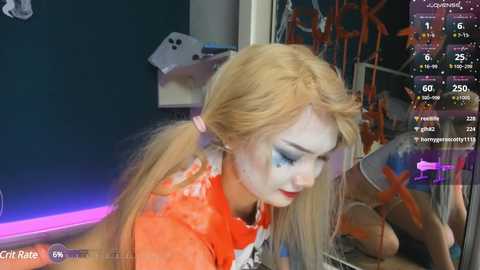 Media: Video of a young woman with fair skin and long blonde hair in a red and white striped top, wearing white face paint and blue eyeshadow, standing in front of a dark green wall with a mirror reflecting her and a digital screen displaying Twitch chat.