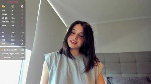 Media: Video of a young woman with straight black hair, light skin, and a smile, wearing a light blue sleeveless top, standing in a modern bedroom with a grey upholstered headboard.