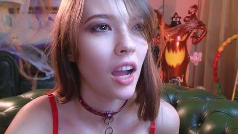 Media: A video of a young woman with straight, light brown hair and a red choker, wearing a red spaghetti strap top, in a dimly lit, colorful, cluttered room.