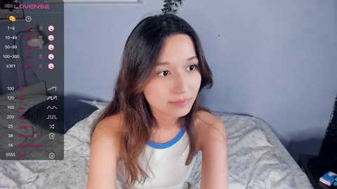 Media: Video of a young Asian woman with long, wavy brown hair, wearing a white tank top, sitting on a bed with gray sheets, in a dimly lit room.