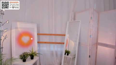 Media: Video of a minimalist, softly lit room with a large white mirror, a vibrant orange sunburst painting, a green potted plant, and white curtains.