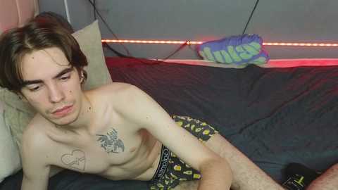 Media: Video of a shirtless, thin, fair-skinned young man with brown hair, lying on a bed with black and yellow patterned pajama bottoms. He has a tattoo on his chest and a pillow with a heart design. Background features a gray wall with red LED lights.