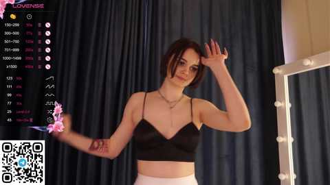 Media: A video of a young woman with short brown hair and fair skin wearing a black crop top and white pants. She stands in front of a dark curtain, holding a pink flower.