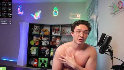 Media: Video of a shirtless, pale-skinned man with glasses, curly hair, and a slim build, standing in a brightly lit room with neon signs, plush toys, and a gaming setup.