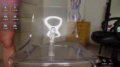 Media: Video of a clear acrylic chair with a large, lit-up white vagina symbol on a wooden floor, featuring a blurred, nude person's leg in the background.