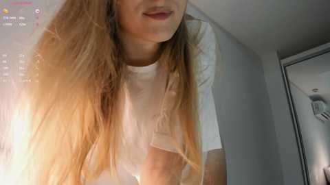 Media: Video of a young woman with long, wavy, light brown hair, wearing a white t-shirt, smiling slightly, in a dimly lit room with a window and a mirror.