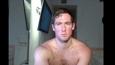 Media: Video of a shirtless white man with short brown hair, wearing white earbuds, standing in a dimly lit room with a flat-screen TV on the left wall and a partially visible door behind him.