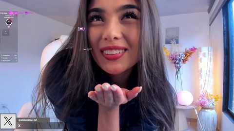 Media: A video of a young woman with long, straight, dark hair, smiling and blowing a kiss, indoors. She wears a dark shirt. The room has white walls, a window, and floral decor.