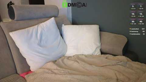 Media: A video of a modern, minimalist bedroom with two large white pillows on a light grey bed, topped with a beige blanket. The wall behind is dark grey. A weather app screen displays temperature and weather icons in the upper right corner.