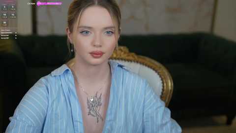 Media: Video of a fair-skinned, blonde-haired woman in a blue striped shirt, seated indoors, wearing a necklace with a heart pendant. Background shows a dark green couch and ornate wooden chair.