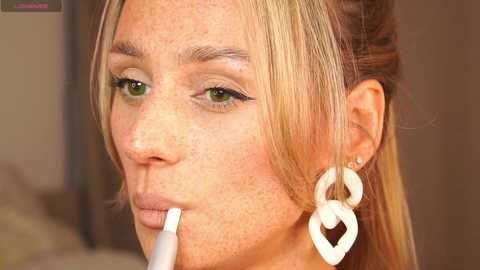 Media: Video of a woman with fair skin, freckles, and green eyes, applying makeup with a white heart-shaped brush. She has blonde hair pulled back, wearing large white heart-shaped earrings. Background is blurred.